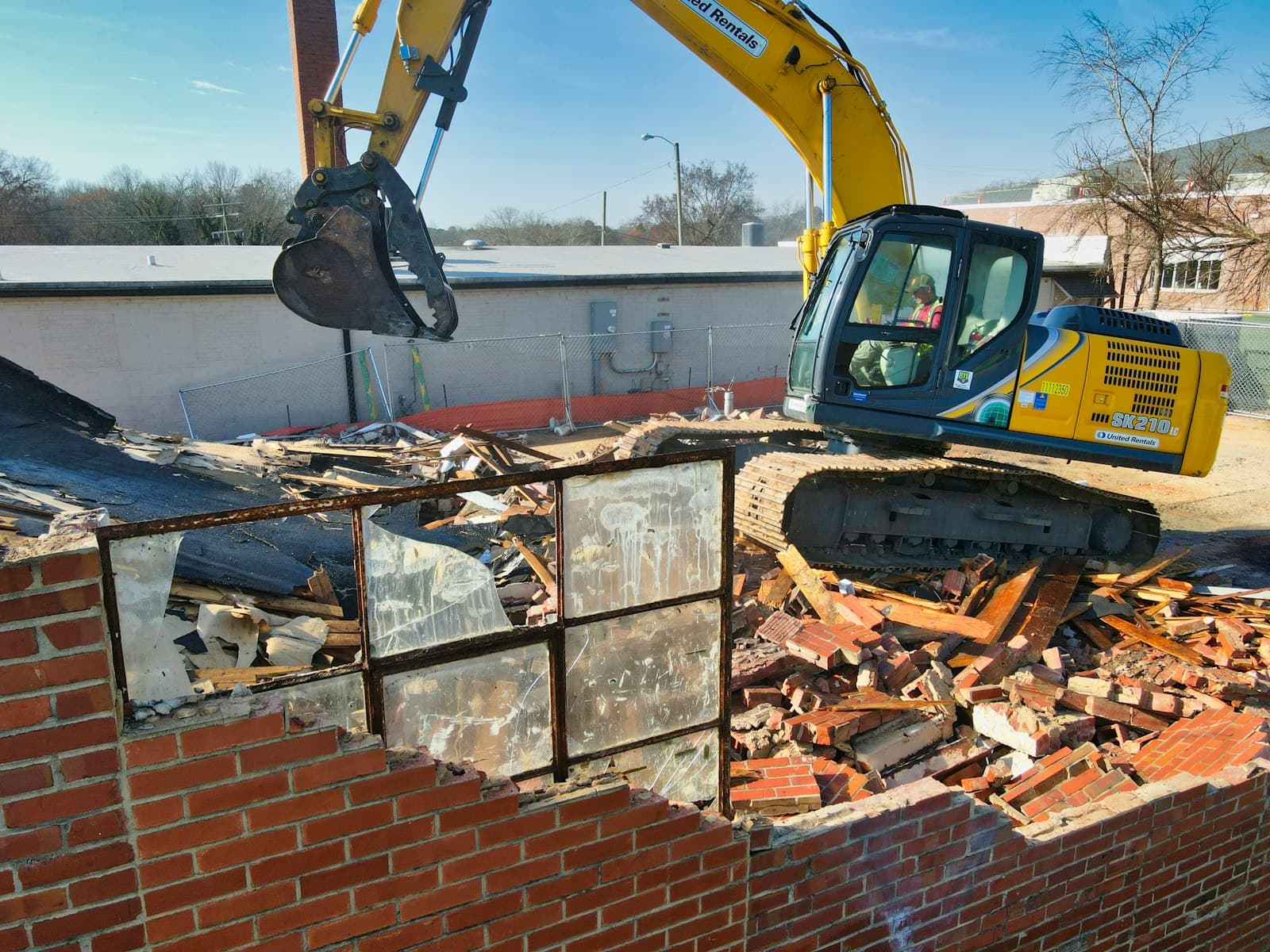 Excavator with operator tearing down a brick building behind site fencing